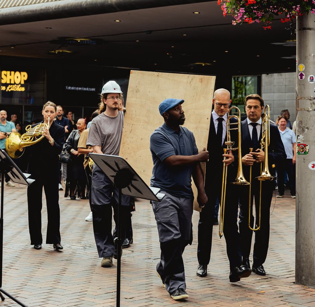 Figuranten met een bouwplaat waar de trombonisten vanachter tevoorschijn komen